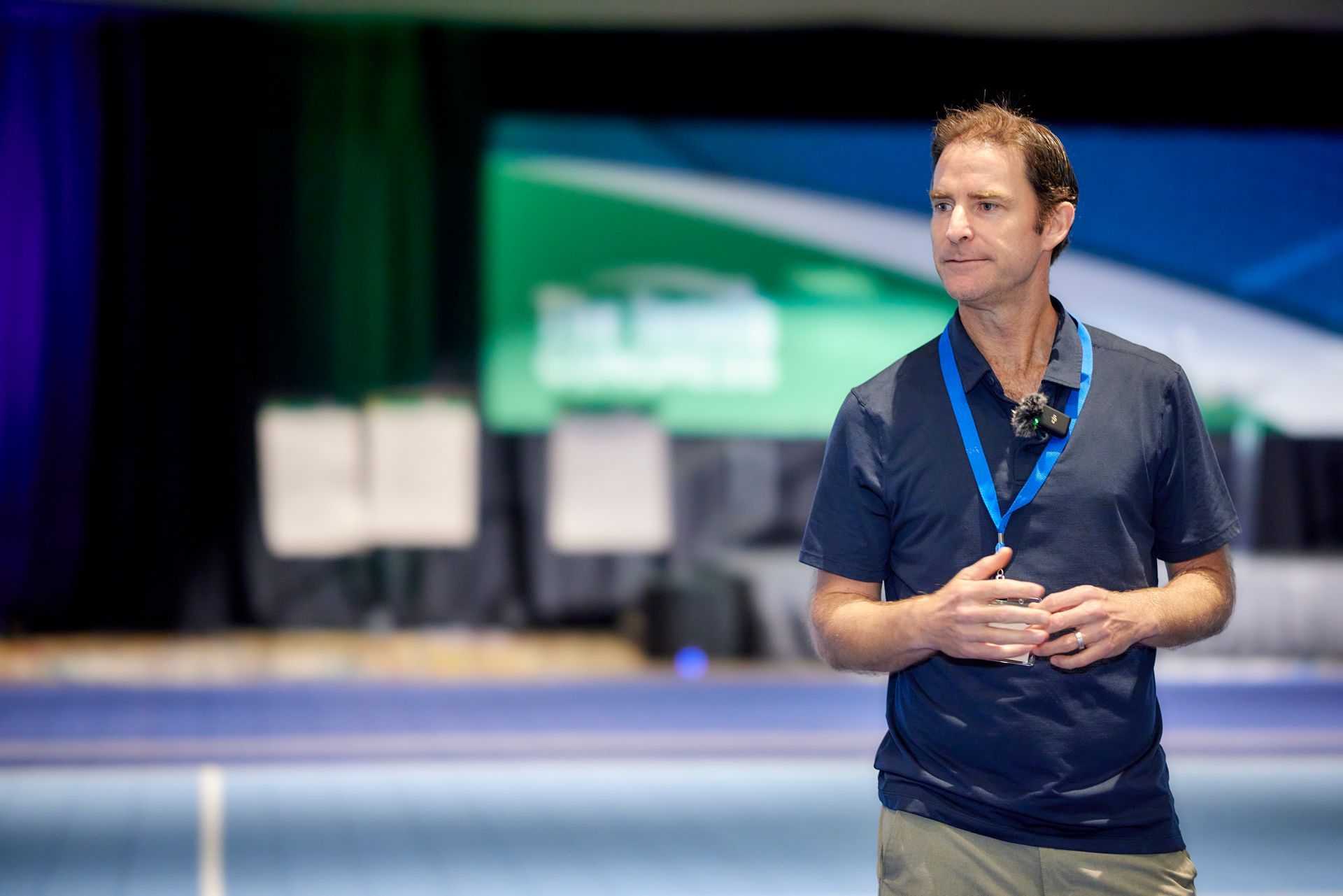 Man in blue shirt, light pants, speaking on stage with backdrop.