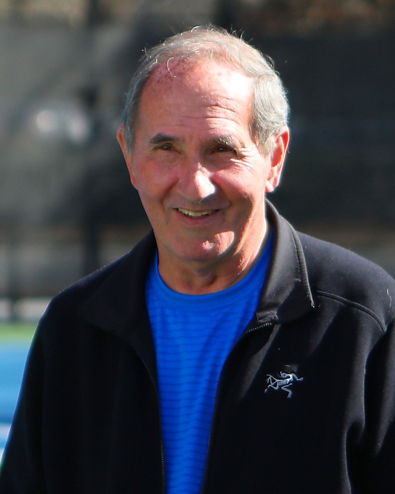 Smiling man in blue shirt and black jacket. Outdoors, sunny day.