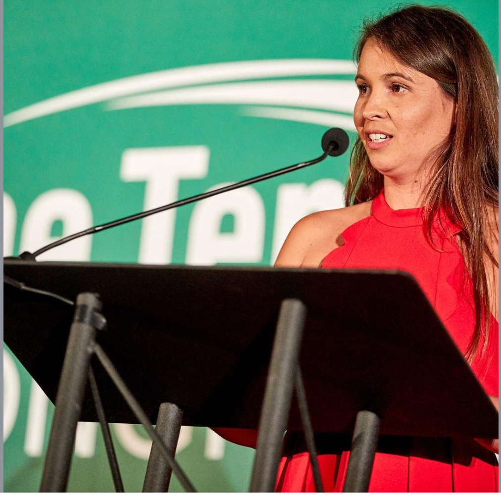Woman in red dress speaking at a podium, with green logo in background.