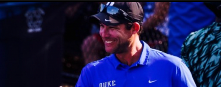 Man in a blue Duke shirt smiles, wearing a black cap with sunglasses on top.