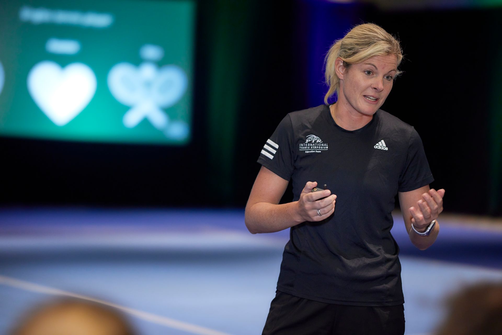 Woman in black Adidas shirt gesturing while speaking in front of a screen with a heart icon.