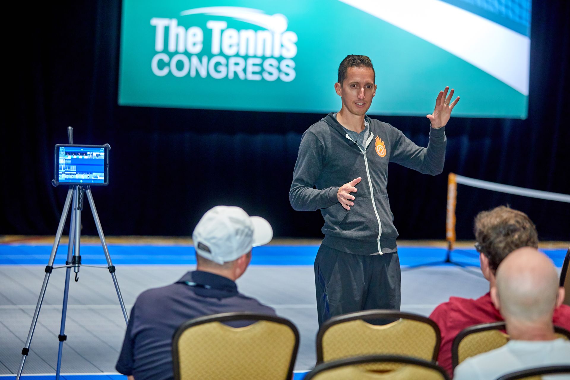 Man giving a tennis coaching presentation. Stage backdrop, audience, tablet on tripod.