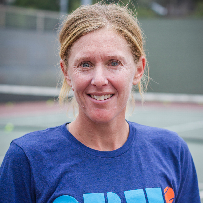 Woman smiling on a tennis court, wearing a blue shirt.