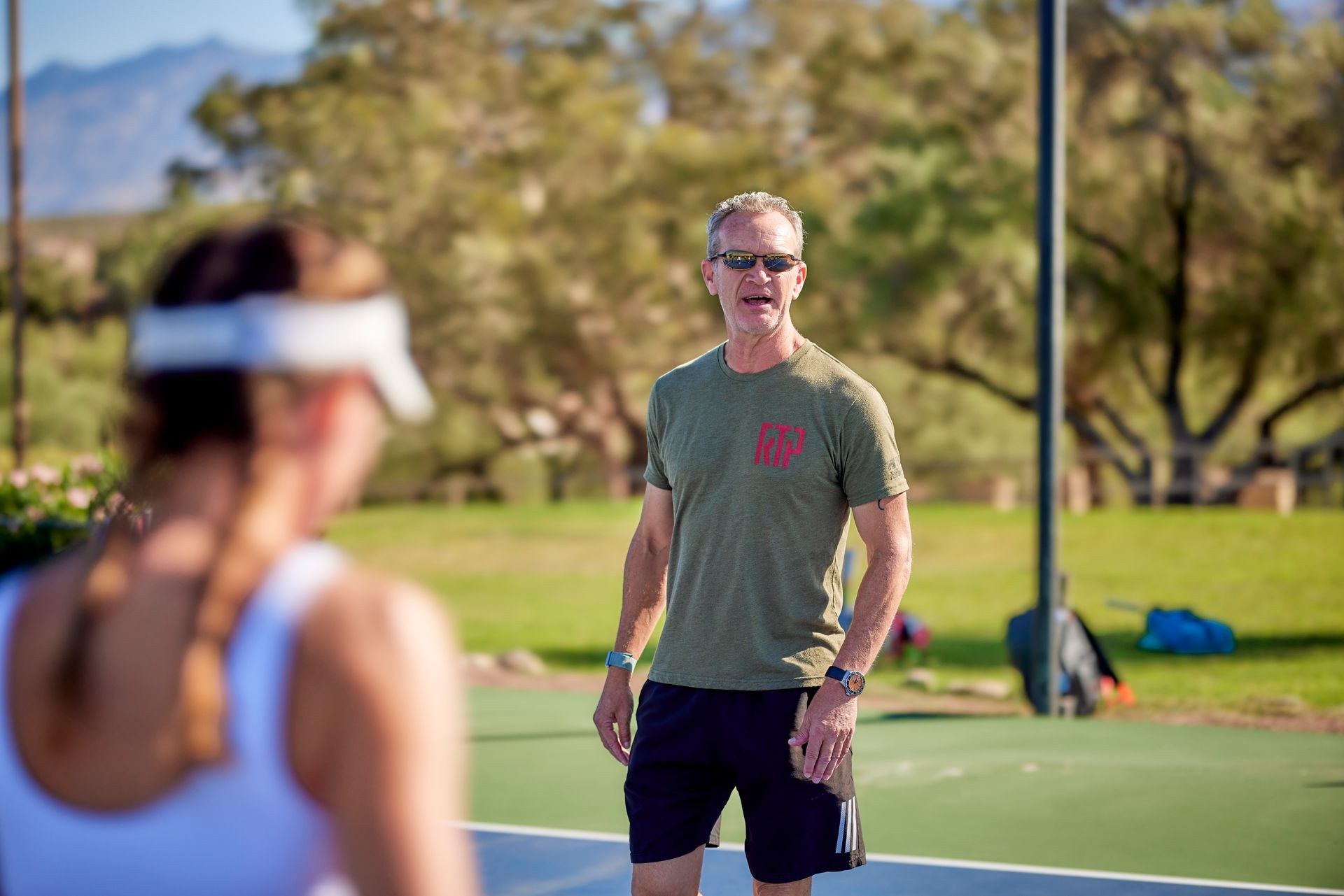 Man in green shirt watches woman playing pickleball outdoors.