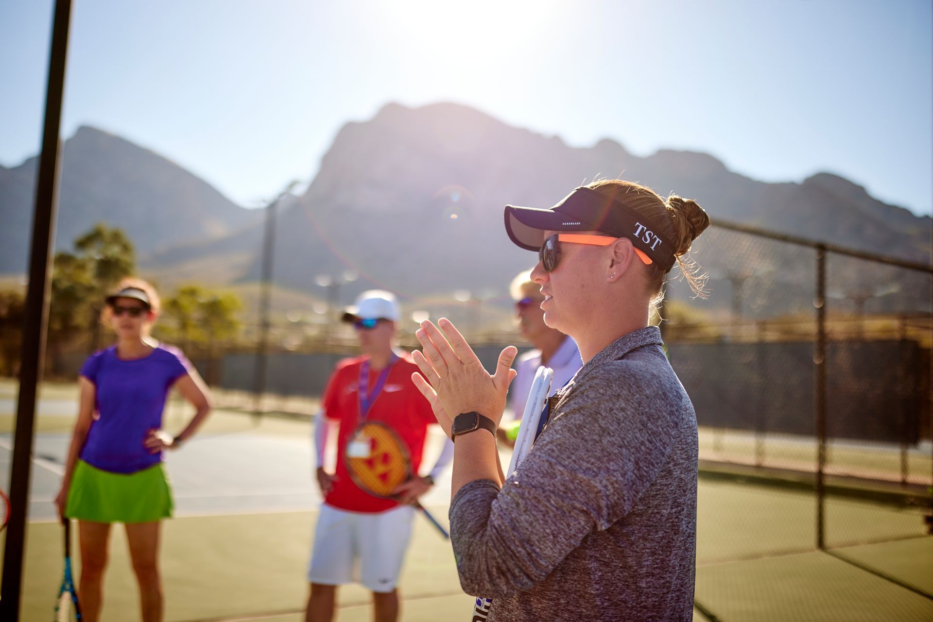 Tennis coach clapping, talking to players on a court, mountains in background.