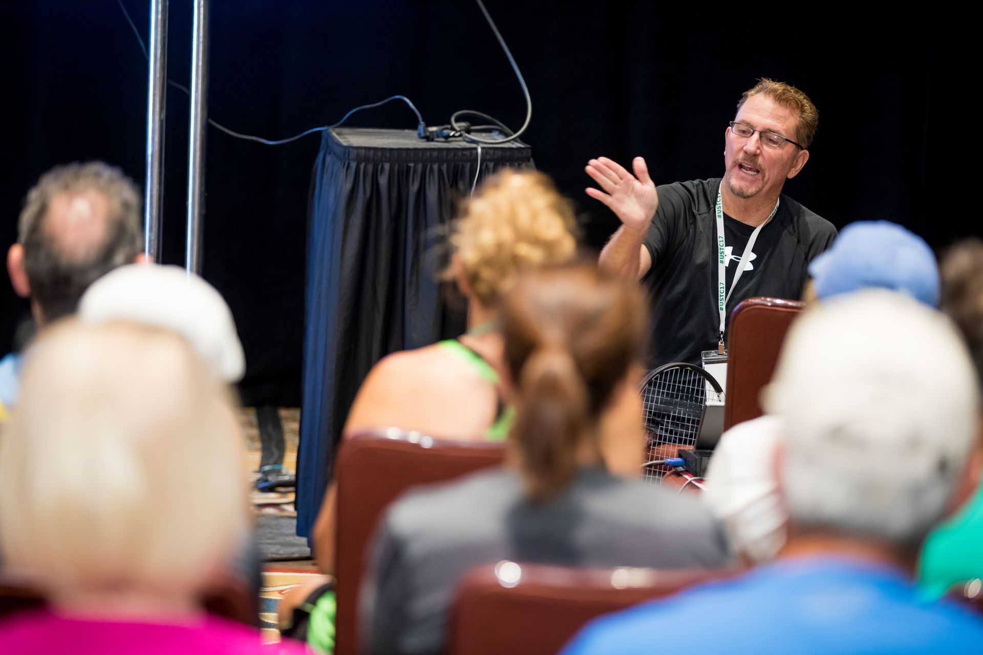 Man gesturing to audience at a presentation. Stage setting, various people in seats.