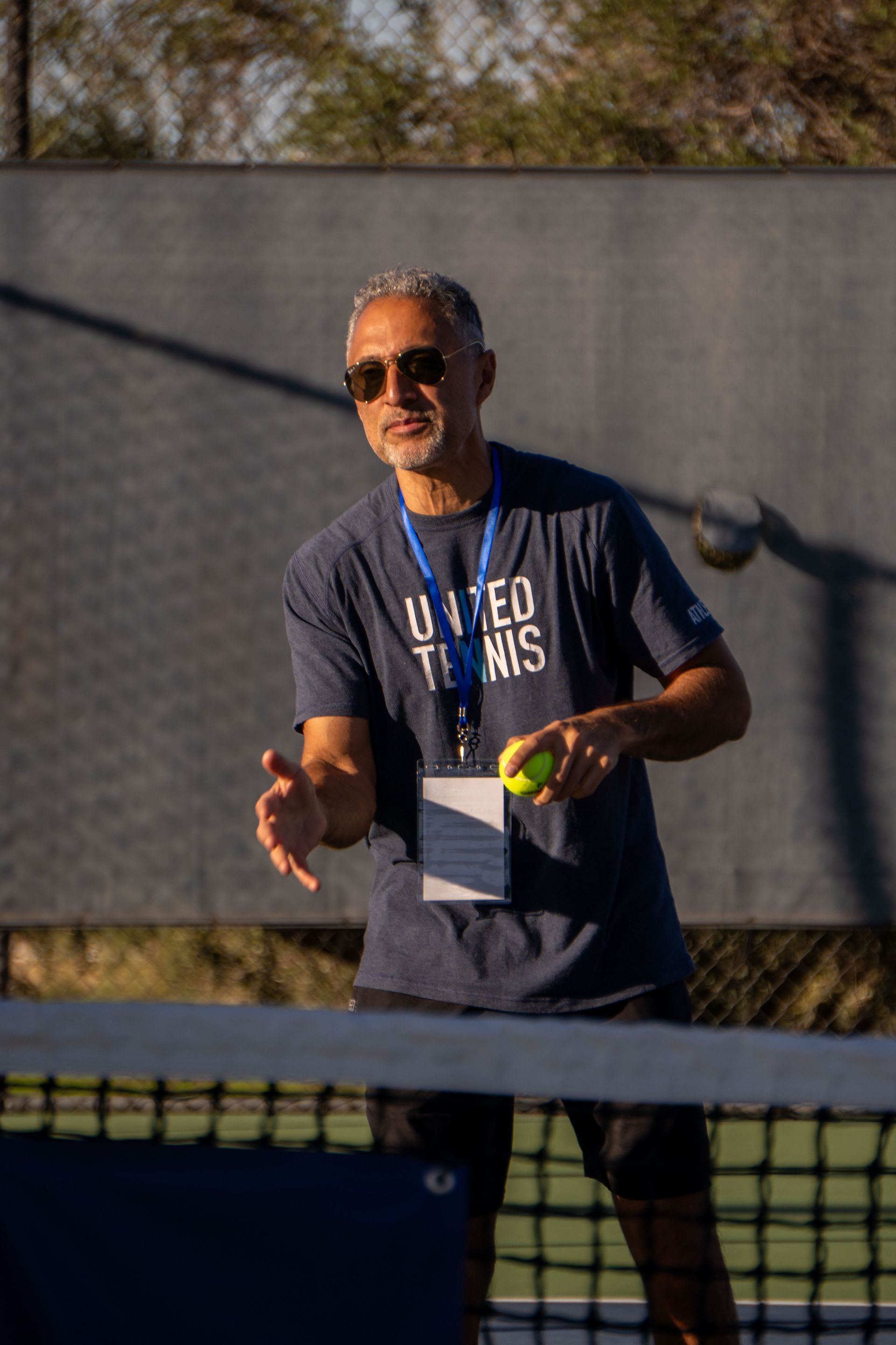 Tennis coach in sunglasses holds a ball, standing on court. Wearing a navy shirt, and a lanyard.