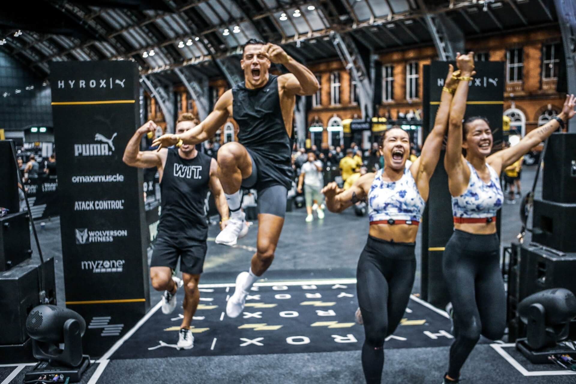 A group of people are jumping in the air at the finish line of a HYROX race event.