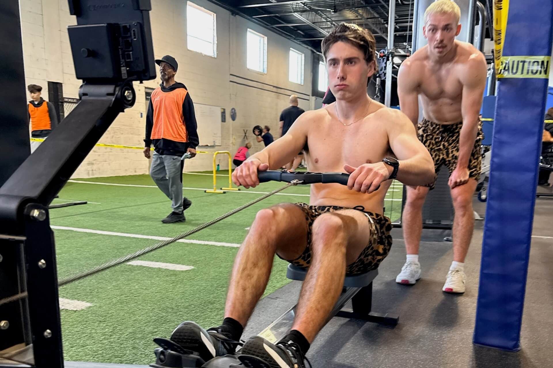 HYROX Athlete on rowing machine in gym, with another athlete standing behind him.
