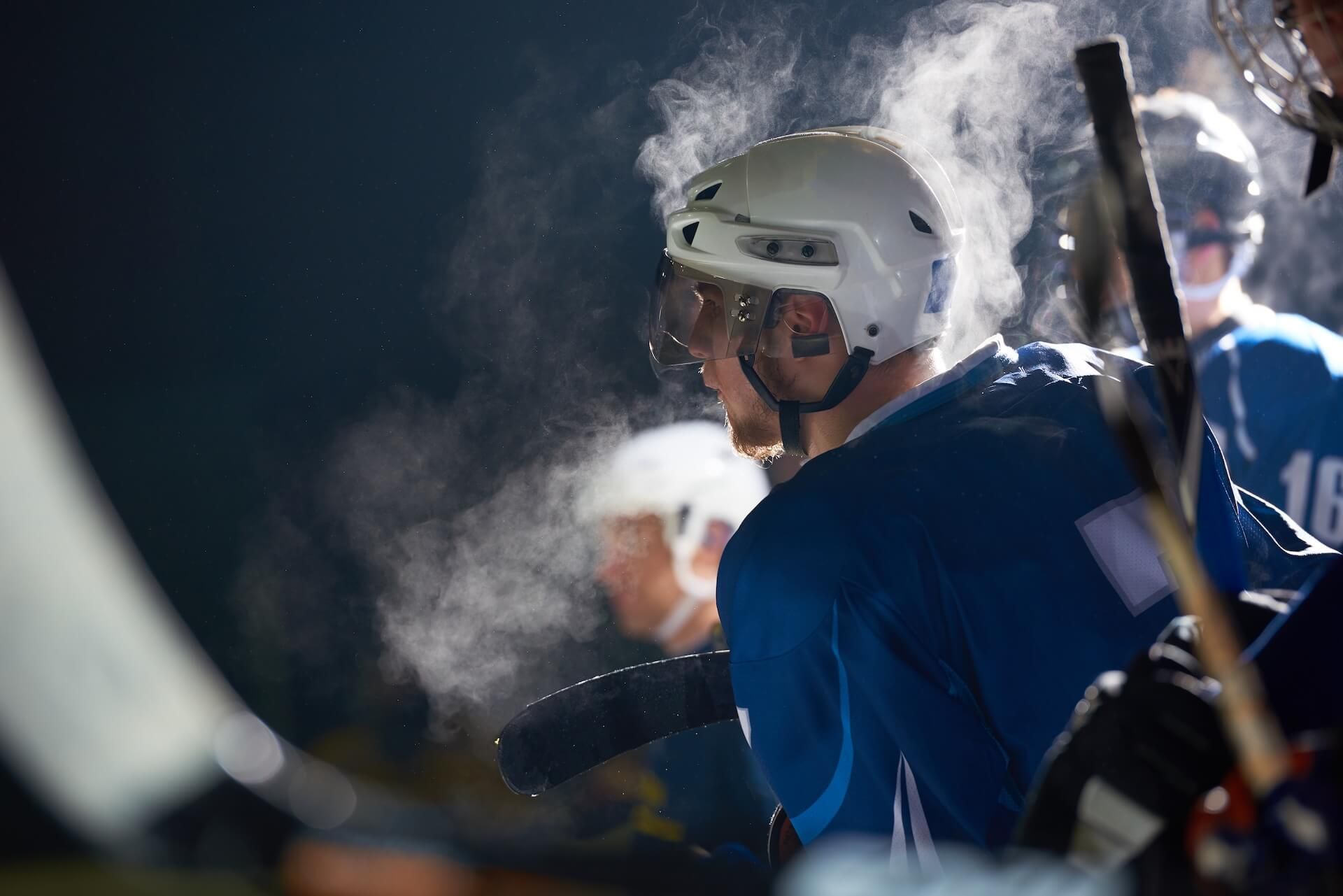 Ice hockey player in blue jersey and white helmet on the bench, breath visible in the cold air