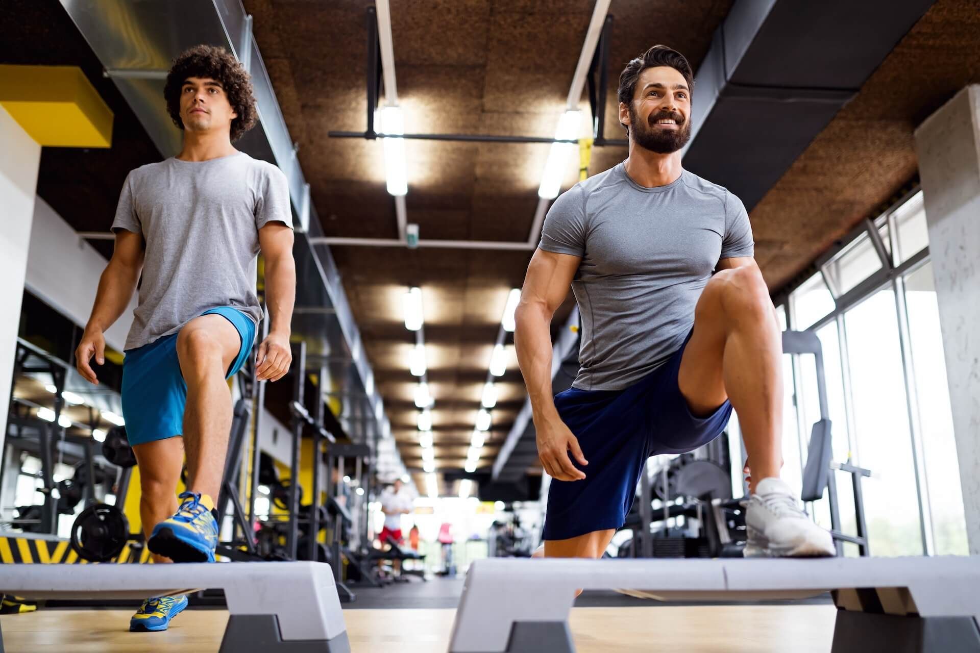 Two people doing knee raises on step platforms in a gym, with bright overhead lights and exercise equipment.