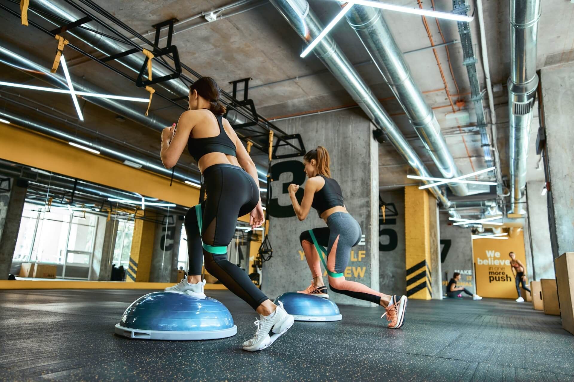 Two people doing balance exercises on blue BOSU balls in a gym with industrial lighting