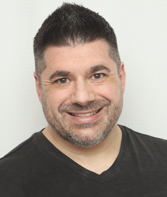 Smiling man with short dark hair and a black V-neck shirt against a light background
