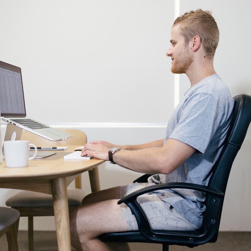 A Man Is Sitting In A Chair At A Desk With A Laptop On It — Sea & Sand Chiropractic In Toukley, NSW