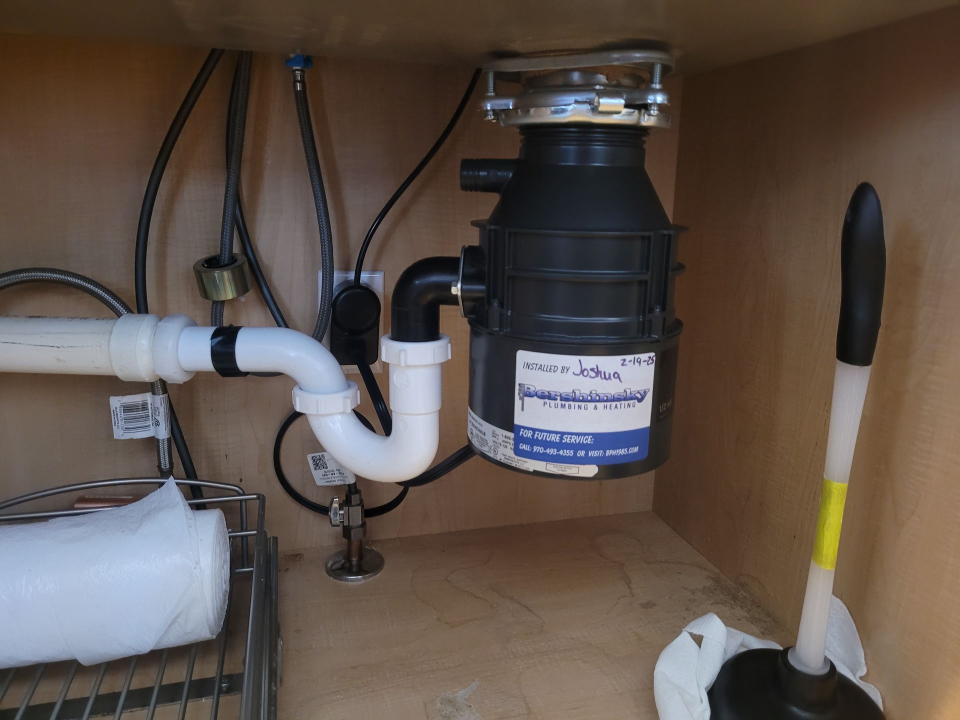 Under-sink cabinet with plumbing pipes, garbage disposal, and a toilet brush on the right