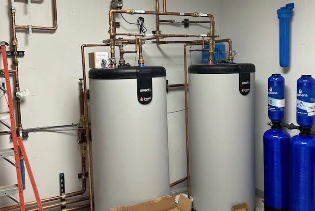 Two large gray water tanks with copper piping in a utility room beside blue water bottles