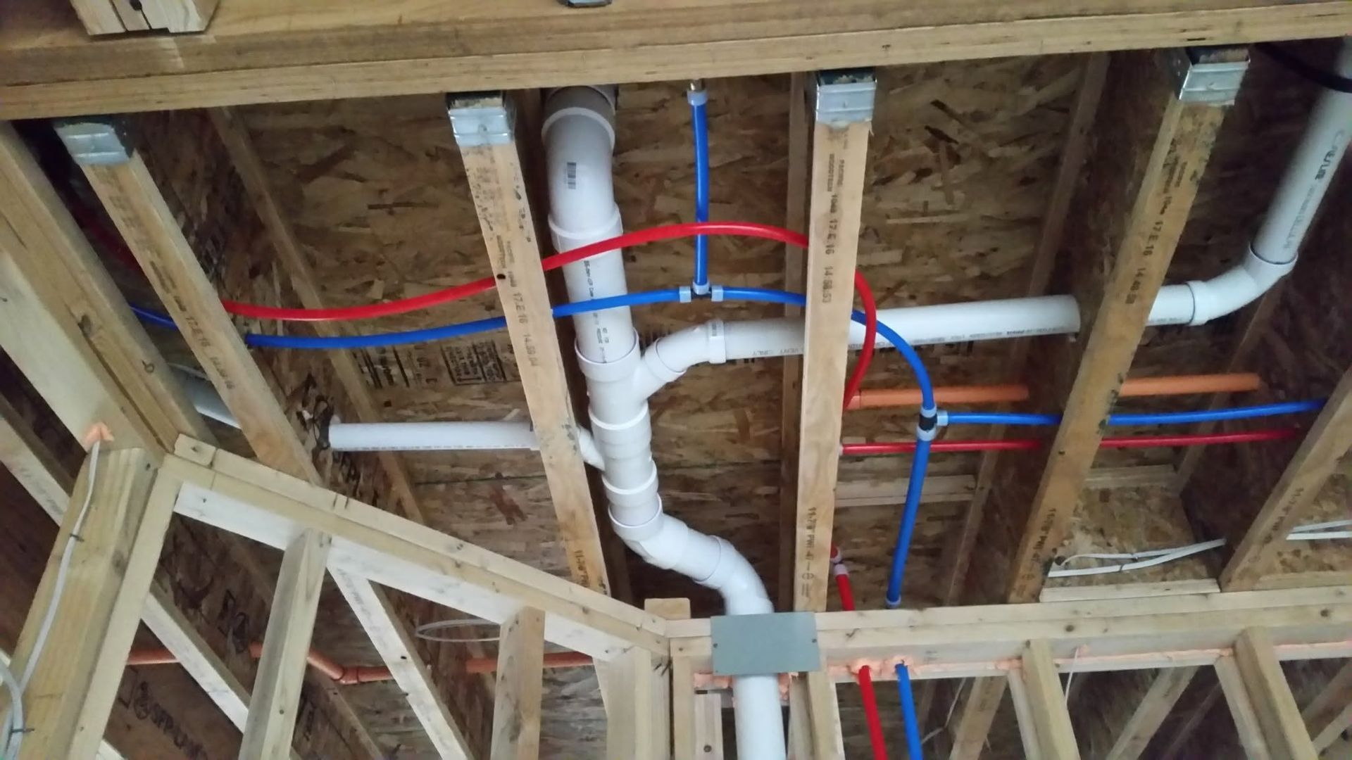 Exposed ceiling framing with white PVC pipes and red and blue PEX plumbing lines