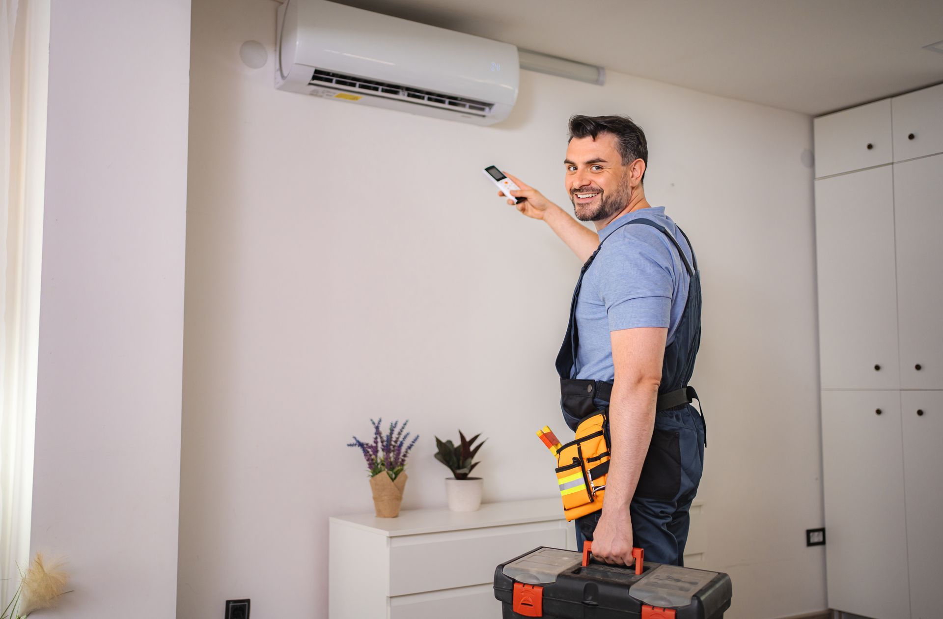 Technician standing on a toolbox, pointing a remote at a wall-mounted air conditioner in a room