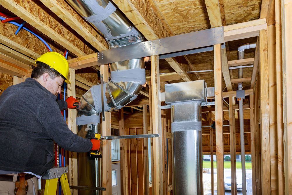 Worker in yellow hard hat installing metal ductwork in a framed building under construction