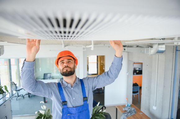 Worker in blue overalls and orange hard hat inspecting a ceiling vent in a bright room