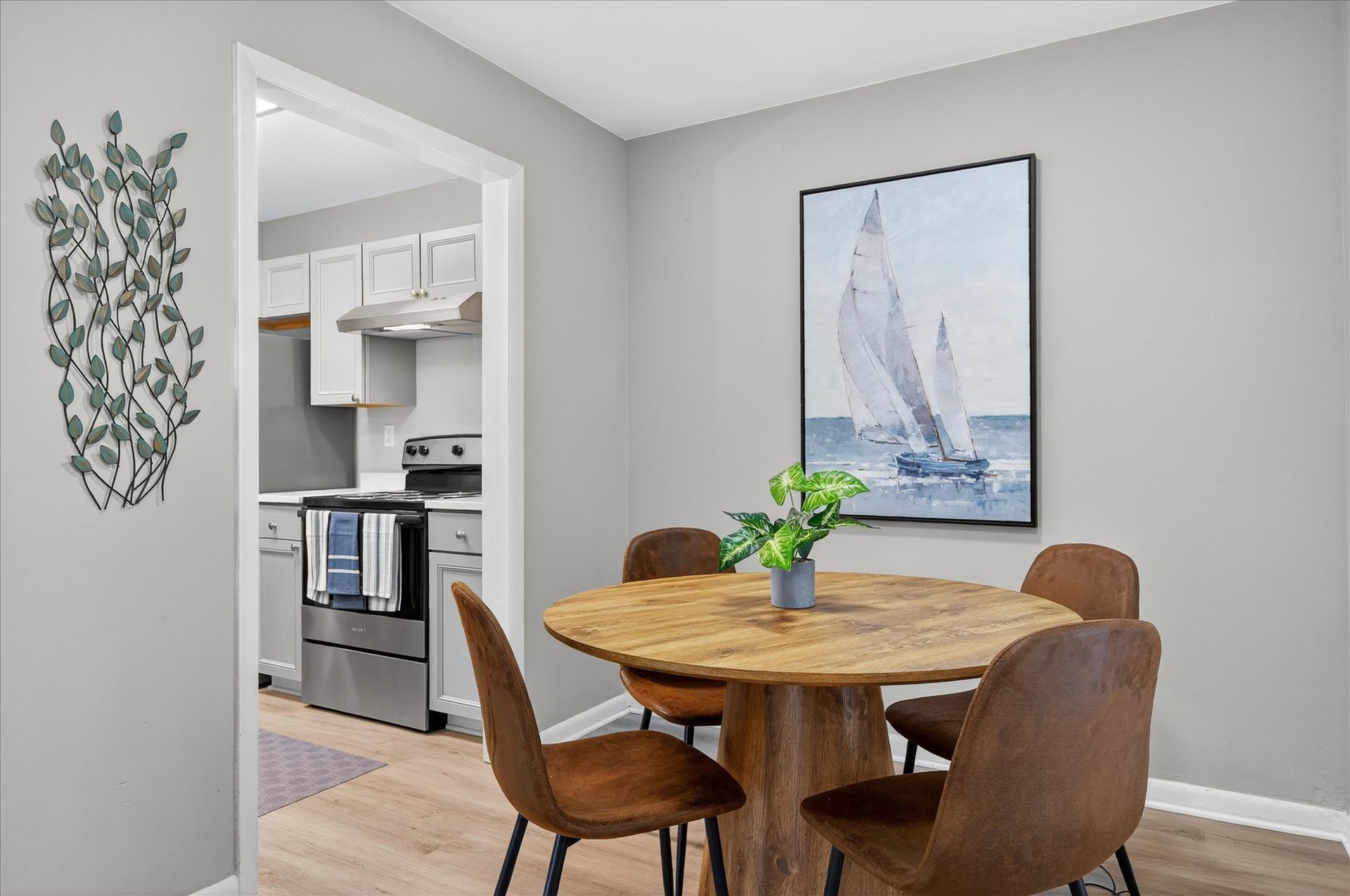 Dining area with round wood table, brown chairs, blue sailboat art, and view into a kitchen.