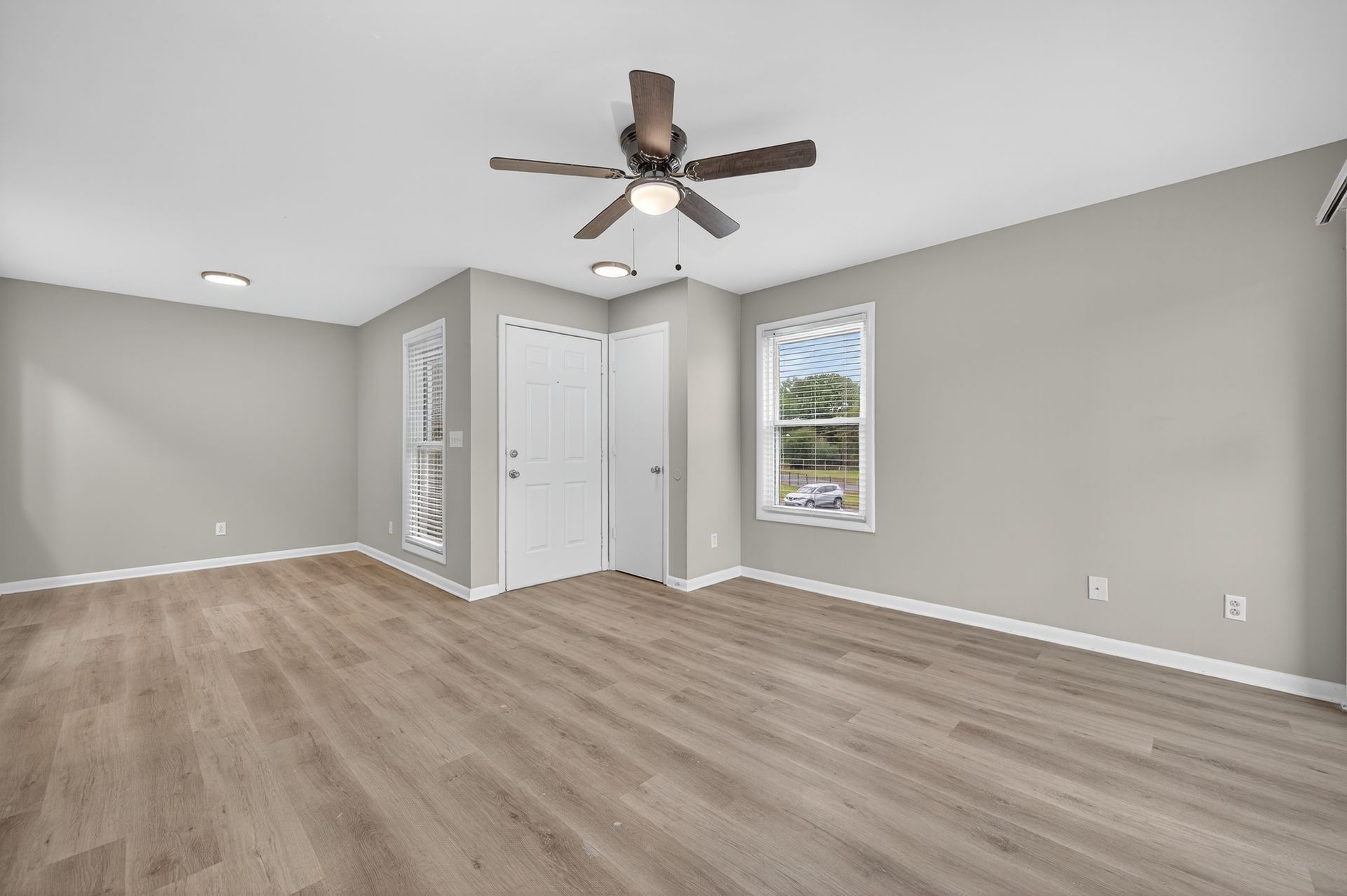Empty gray bedroom with wood flooring, ceiling fan, closet doors, and a window.