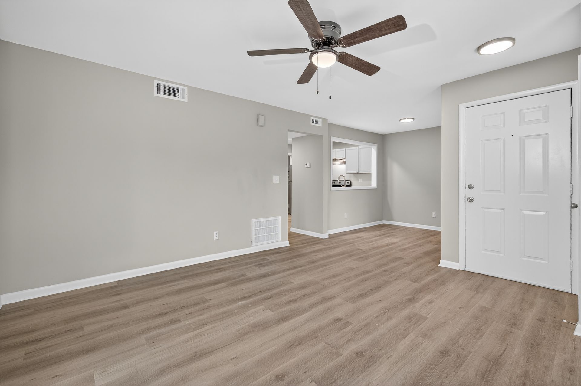 Empty modern living room with gray walls, wood floor, ceiling fan, and a doorway to a kitchen area