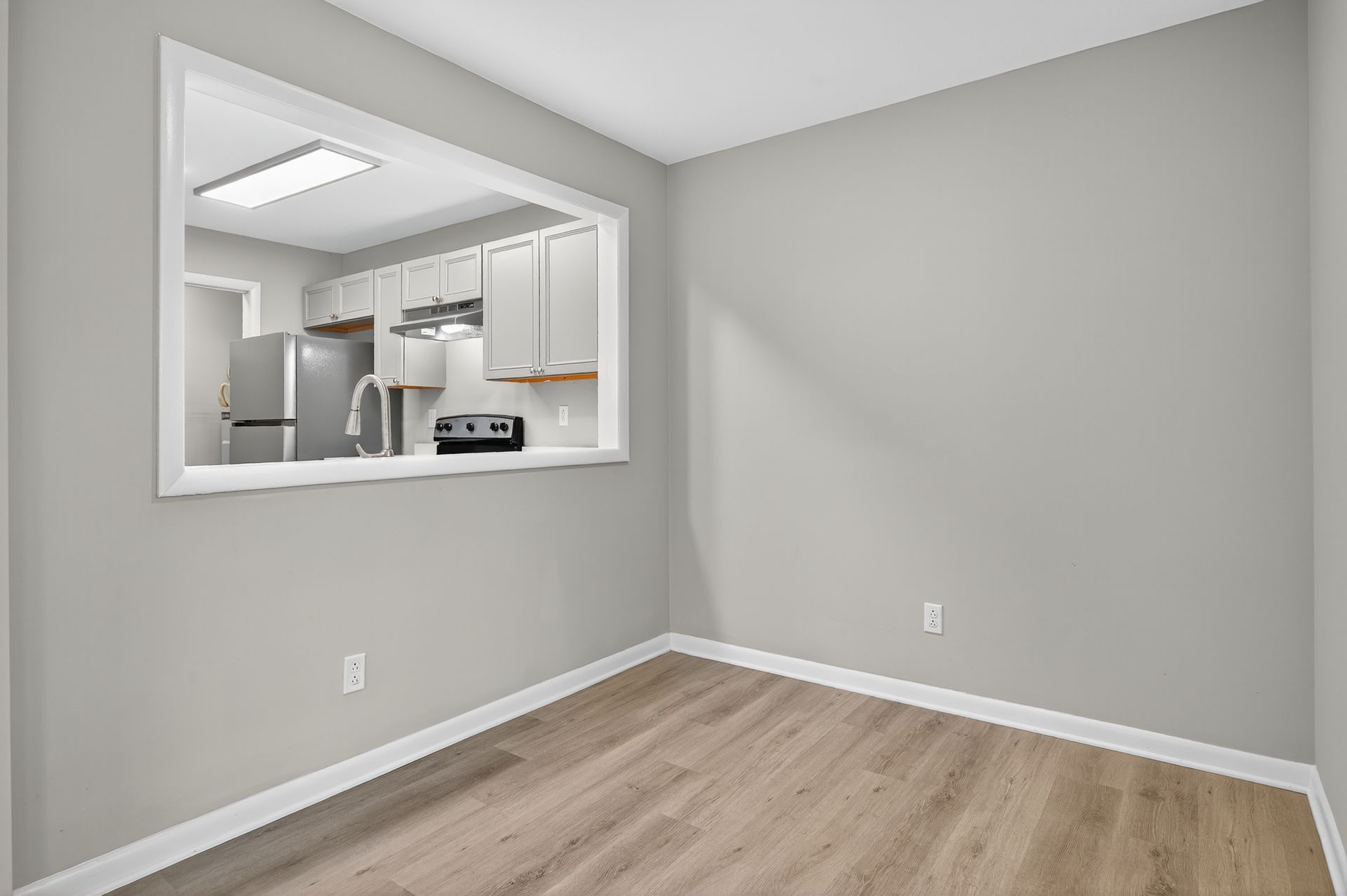 Empty gray room with wood floor and a pass-through window to a bright kitchen