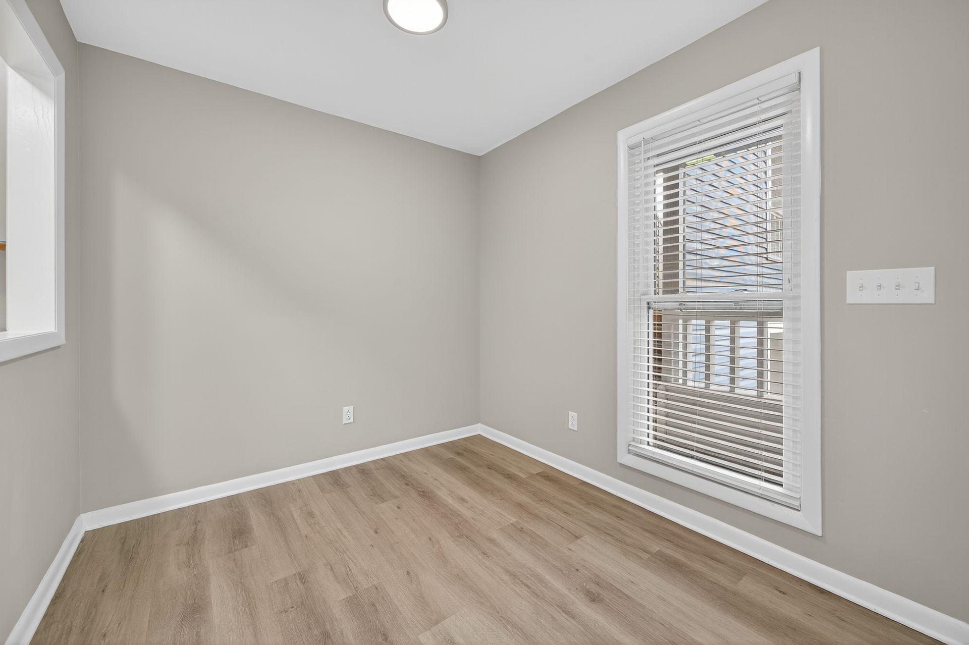 Empty beige room with light wood floor, white trim, and a window with blinds