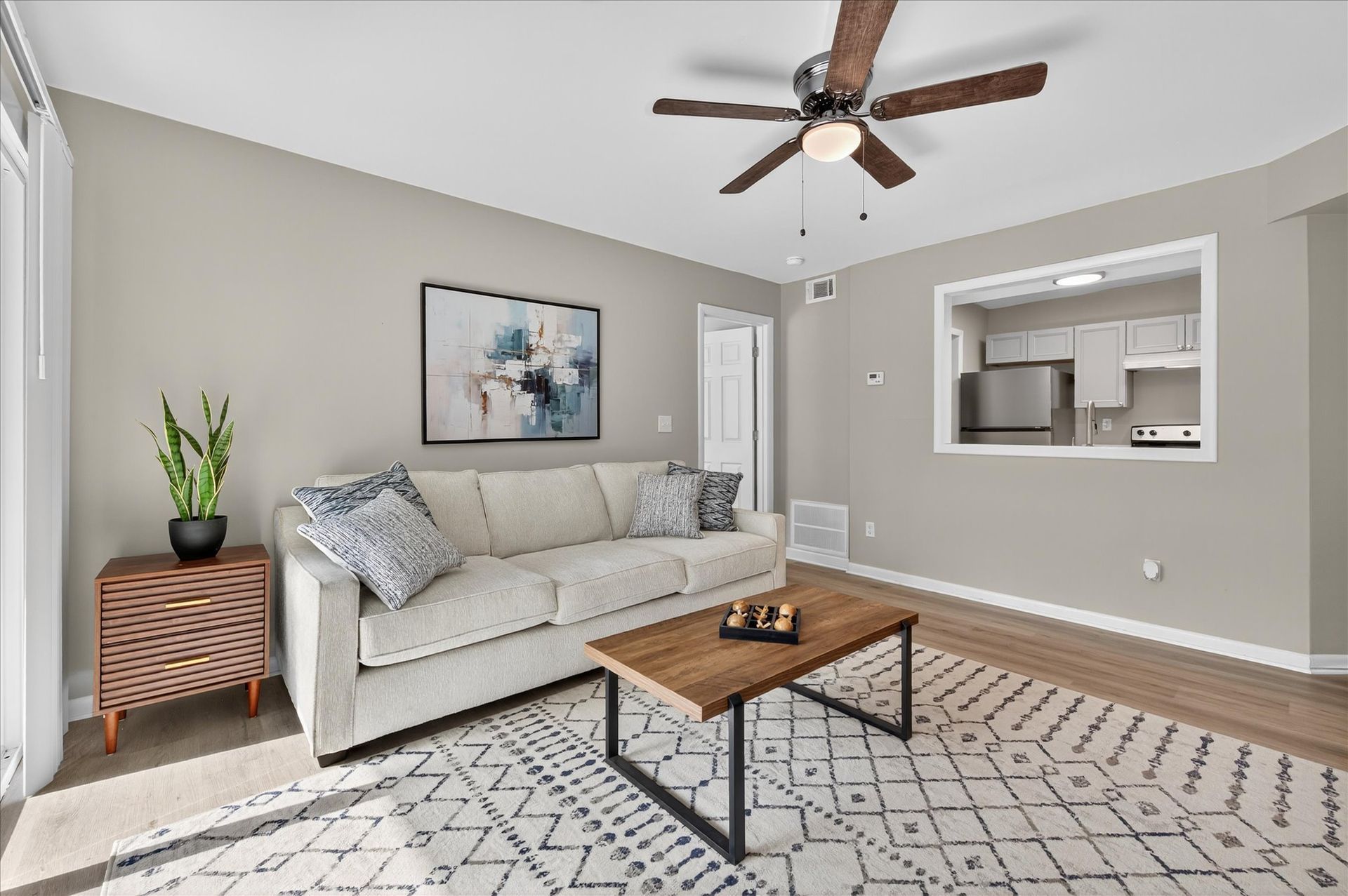 Bright living room with beige sofa, patterned rug, coffee table, ceiling fan, and open kitchen pass-through.