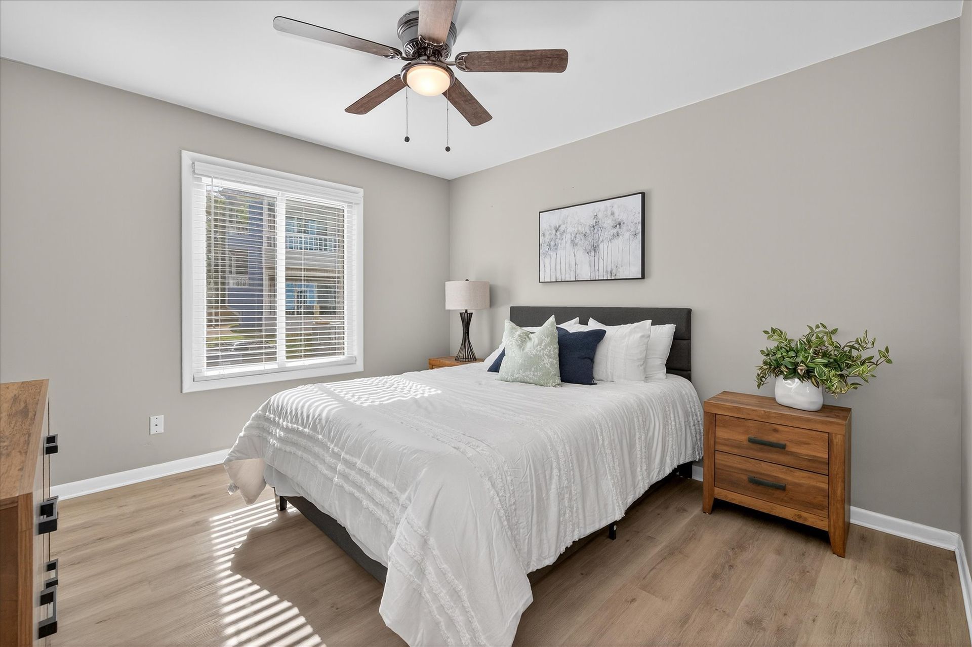Bright bedroom with a bed, ceiling fan, window, lamp, and wooden nightstand with flowers