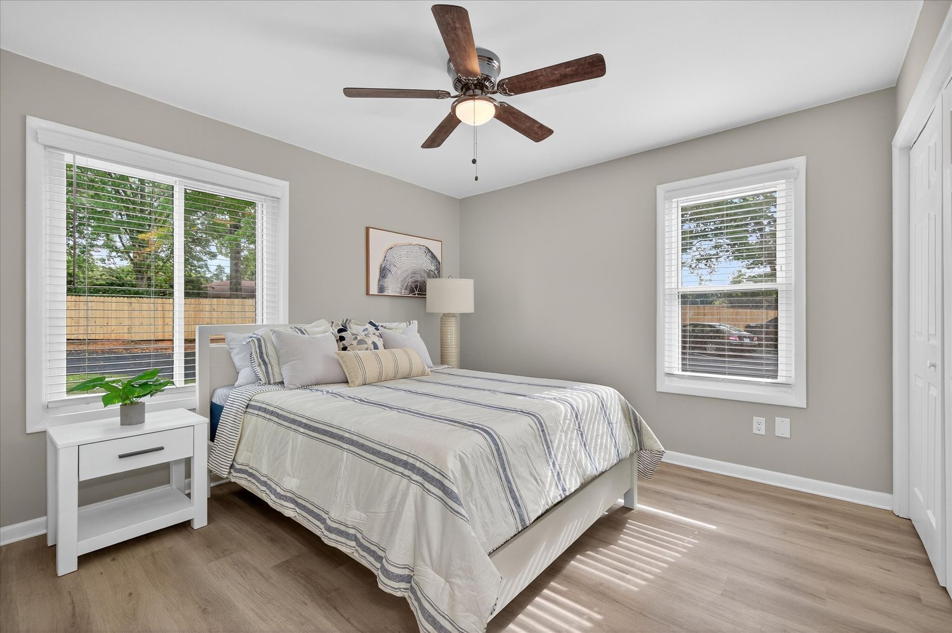 Bright bedroom with a bed, nightstand, lamp, ceiling fan, and two windows with sunlight.