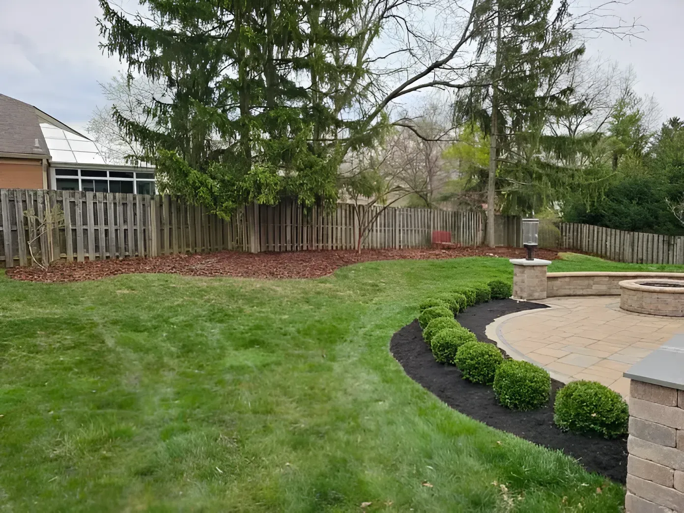 A grassy backyard features a stone patio with a curved garden bed of small, round green bushes and a wooden fence.