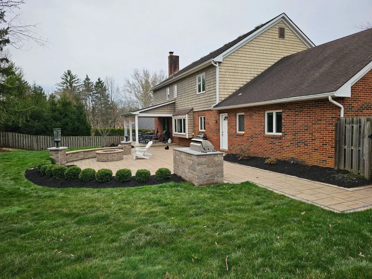 A tan brick patio with a stone fire pit and grill island behind a residential house with tan siding and brick exterior.