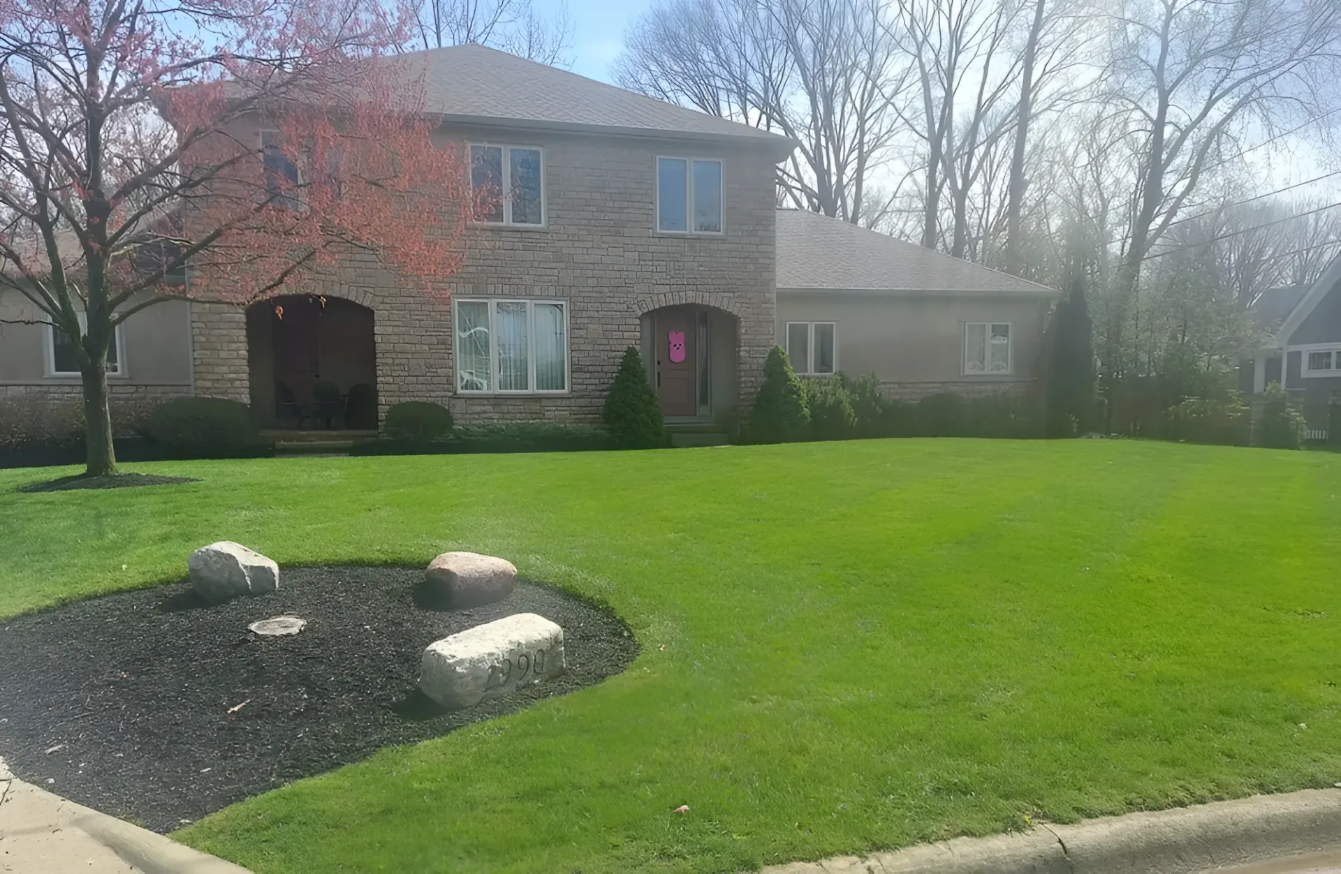 A two-story brick house with a lawn, a mulched garden bed with large rocks, and trees under a clear, bright sky.