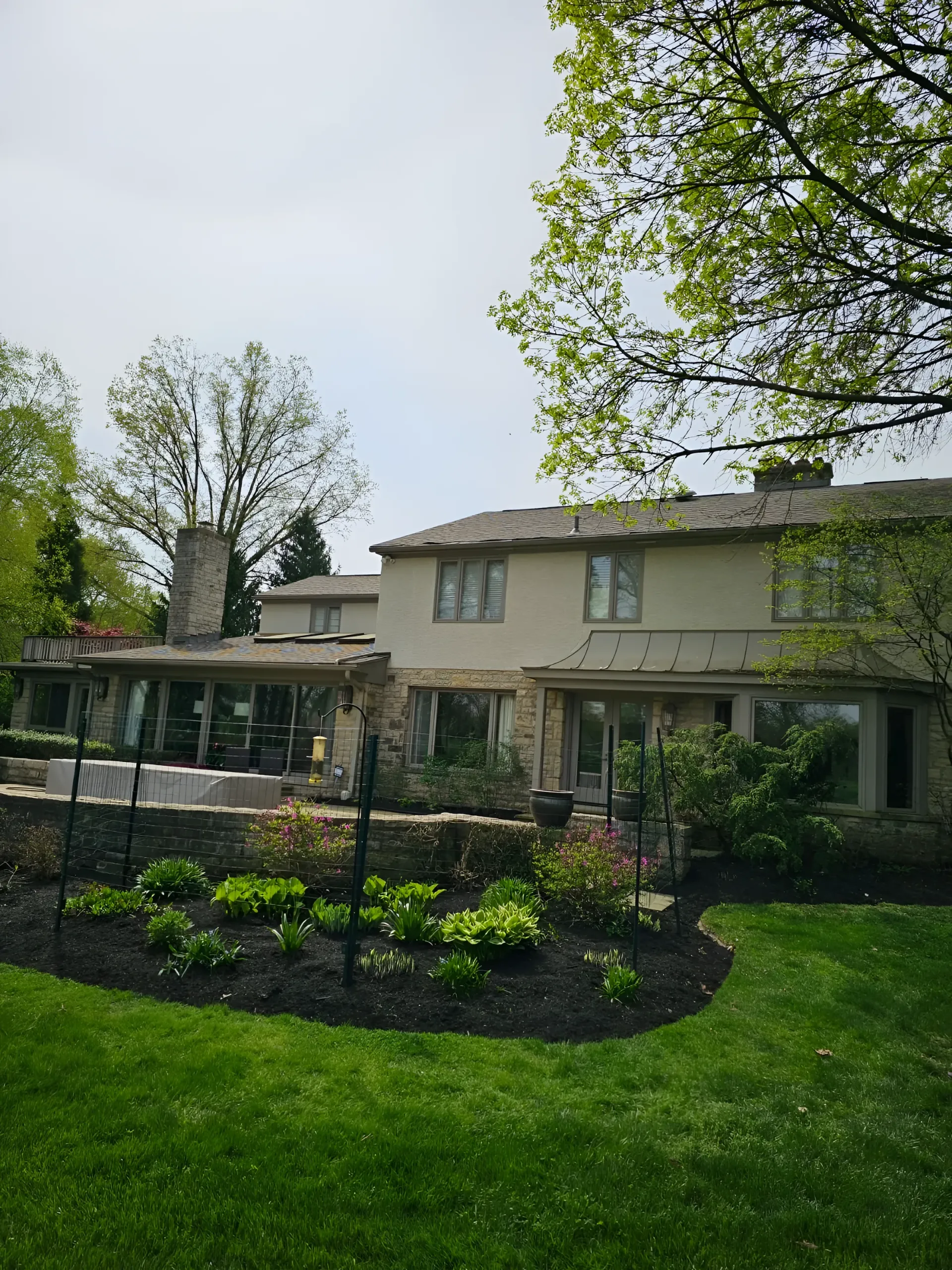 A two-story tan house with a stone patio, garden beds with black mulch, and a green lawn under a partially cloudy sky.