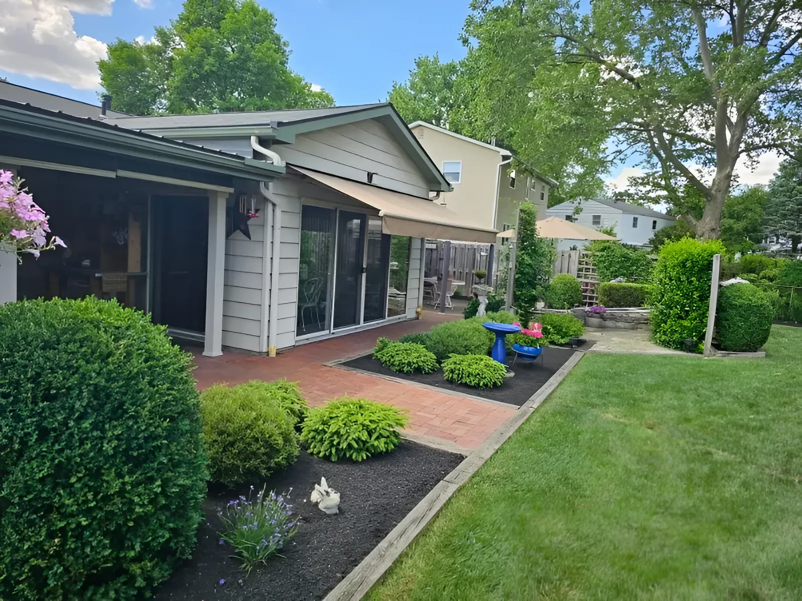 A house with a brick patio, manicured shrubs, and a retractable beige awning, viewed from a grassy backyard on a sunny day.