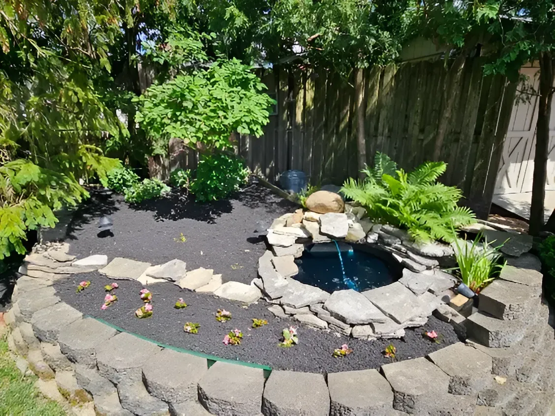 A landscaped backyard garden feature with a circular pond, rock border, and small plants set in dark mulch.