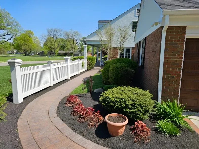 A curved brick walkway leads past a white picket fence, a house, and landscaped bushes in a garden on a sunny day.