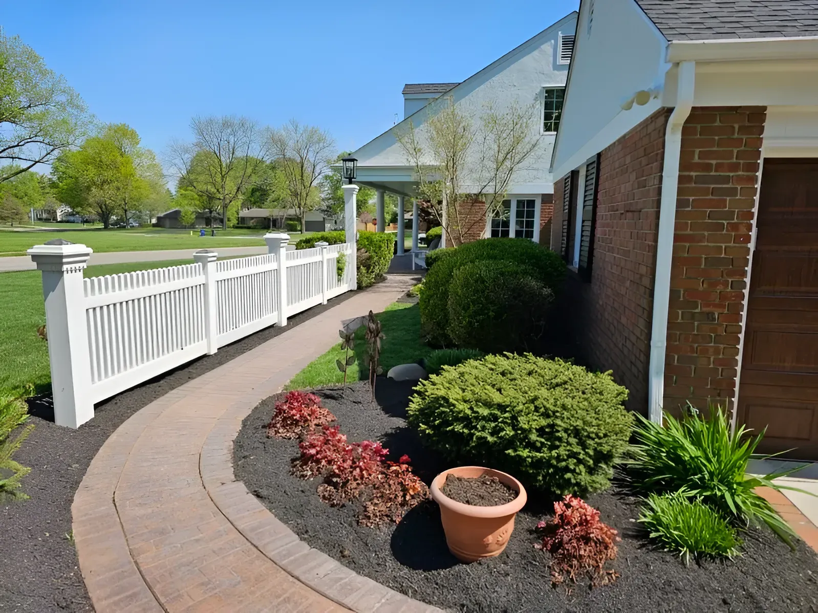 A brick house with a paved walkway, white picket fence, and landscaped garden beds on a sunny day.