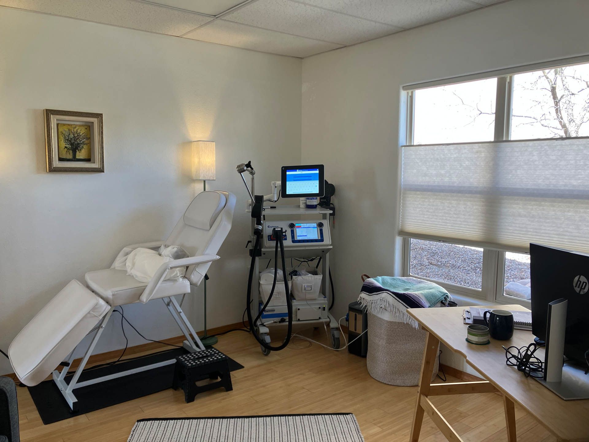Medical exam room with reclined TMS therapy device and chair, monitor, lamp, and desk by a window.