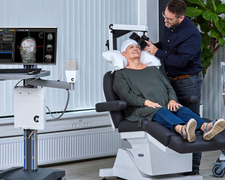 A man is standing next to a woman sitting in a TMS chair being treated for depression.