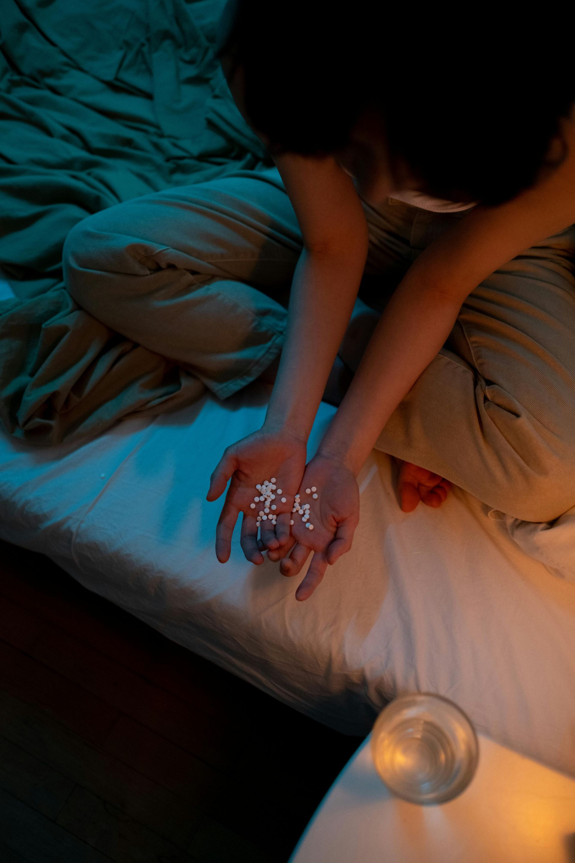 Person holding pills on a bed; a glass of water nearby.