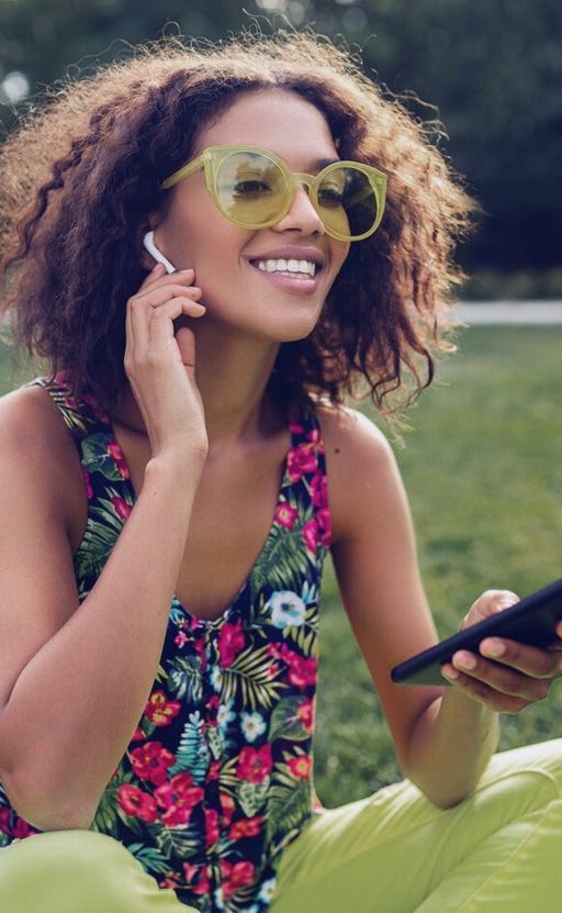 A woman wearing headphones and sunglasses is sitting in the grass using a cell phone.