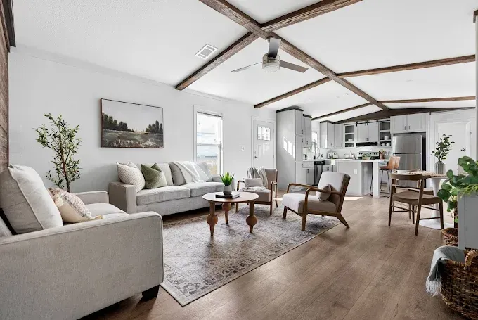 Bright open-plan living room with gray sofas, wood beams, coffee table, and kitchen in the background