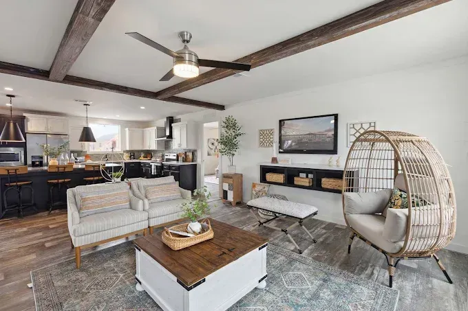 Modern open-concept living room with exposed beams, gray sofa, wicker chair, coffee table, and kitchen in background