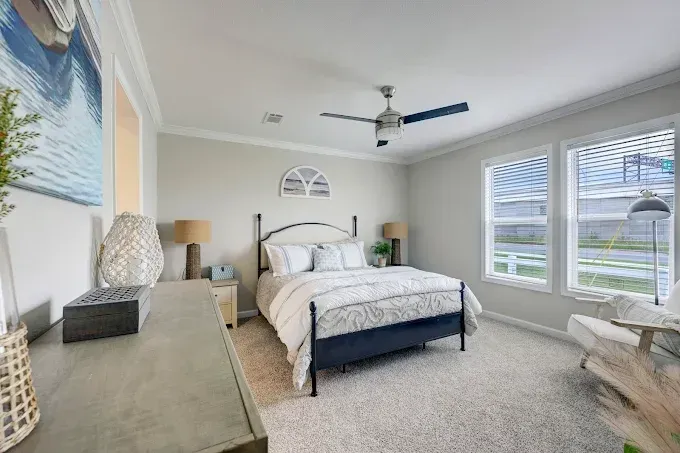 Bright bedroom with a large bed, ceiling fan, light carpet, and windows overlooking a yard