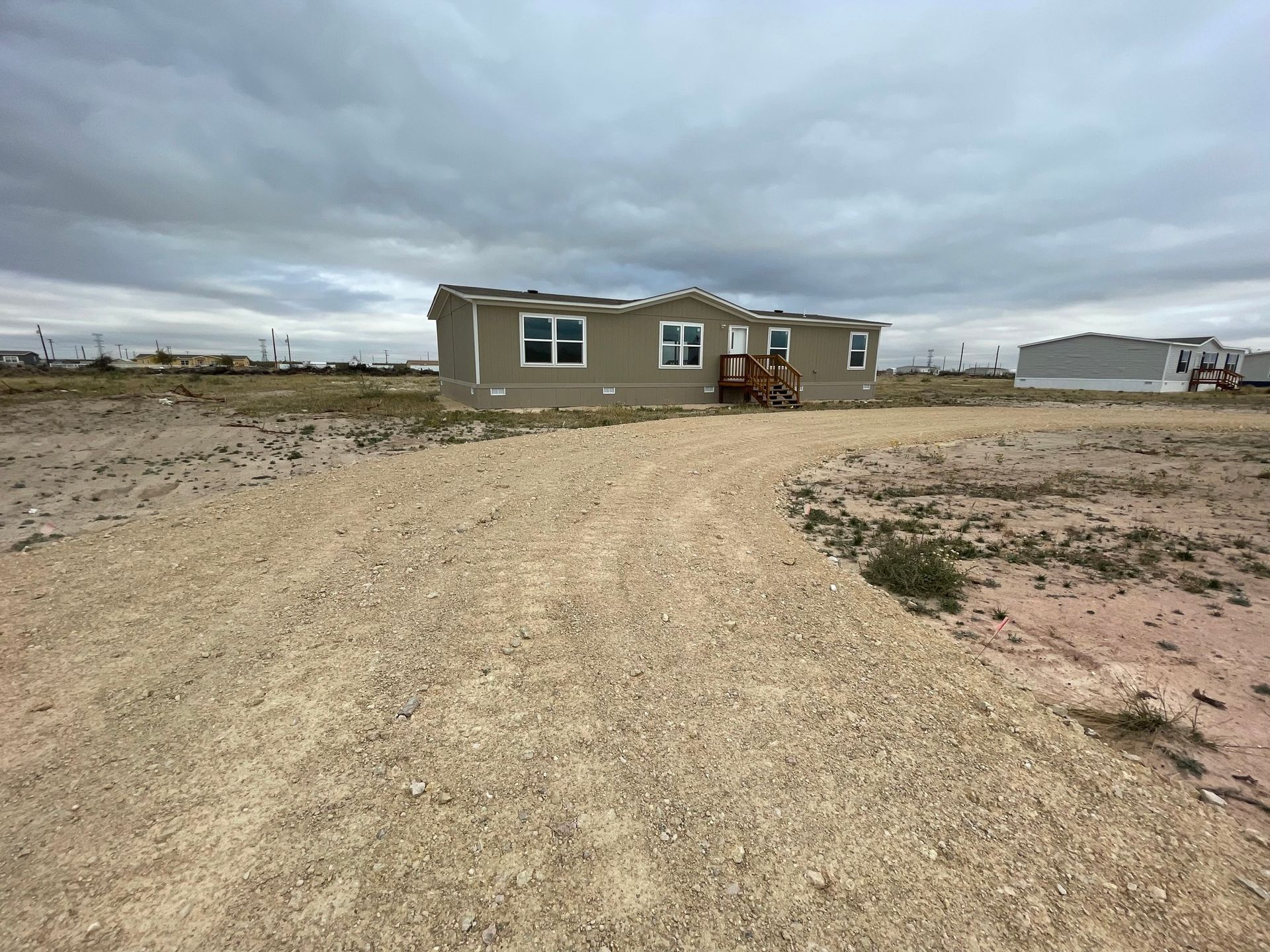 Small house in a barren desert lot under a cloudy sky, with a dirt road leading to the front door.