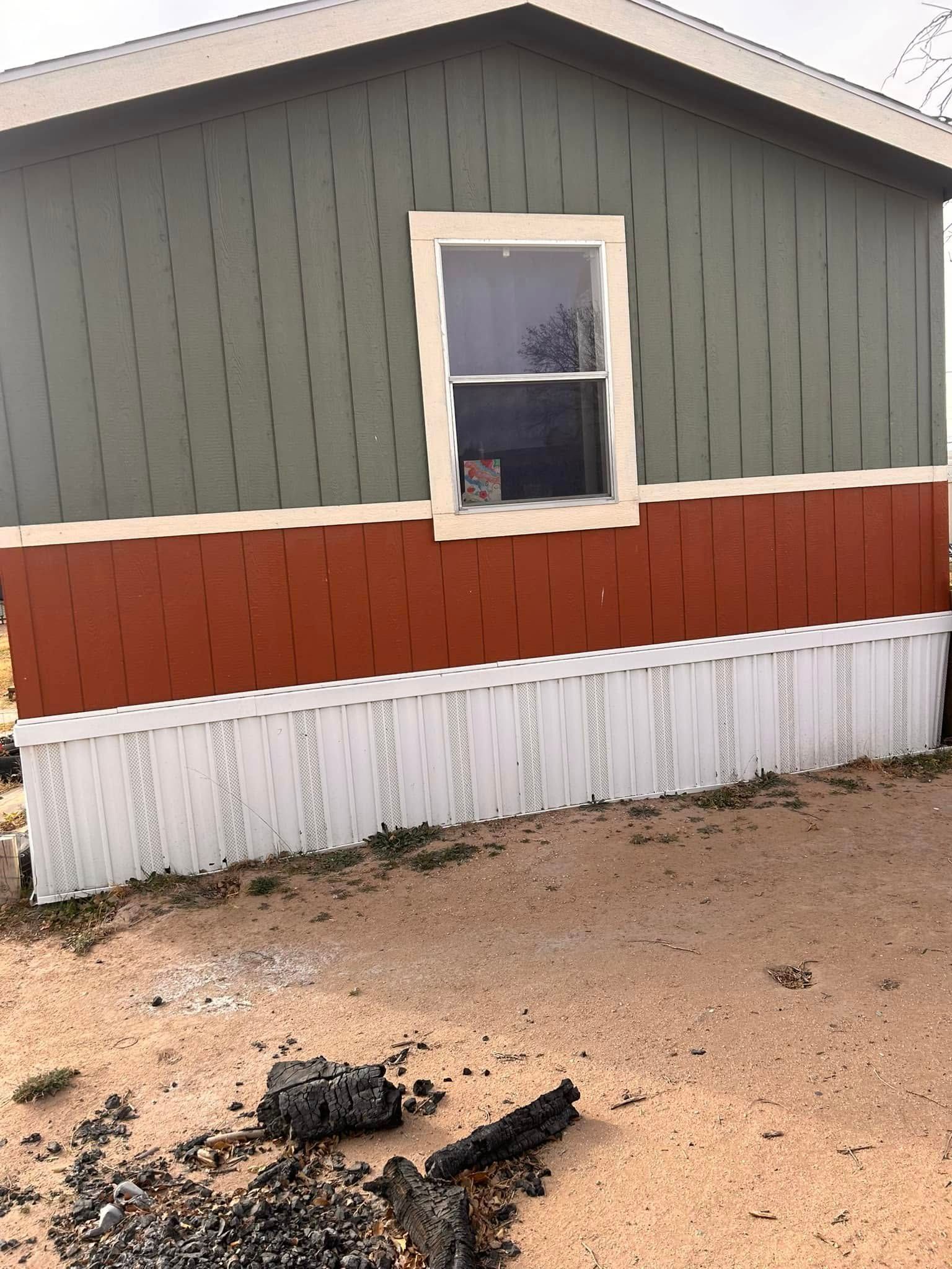 Small house exterior with gray siding, red trim, white skirting, and a charred debris pile in the foreground.