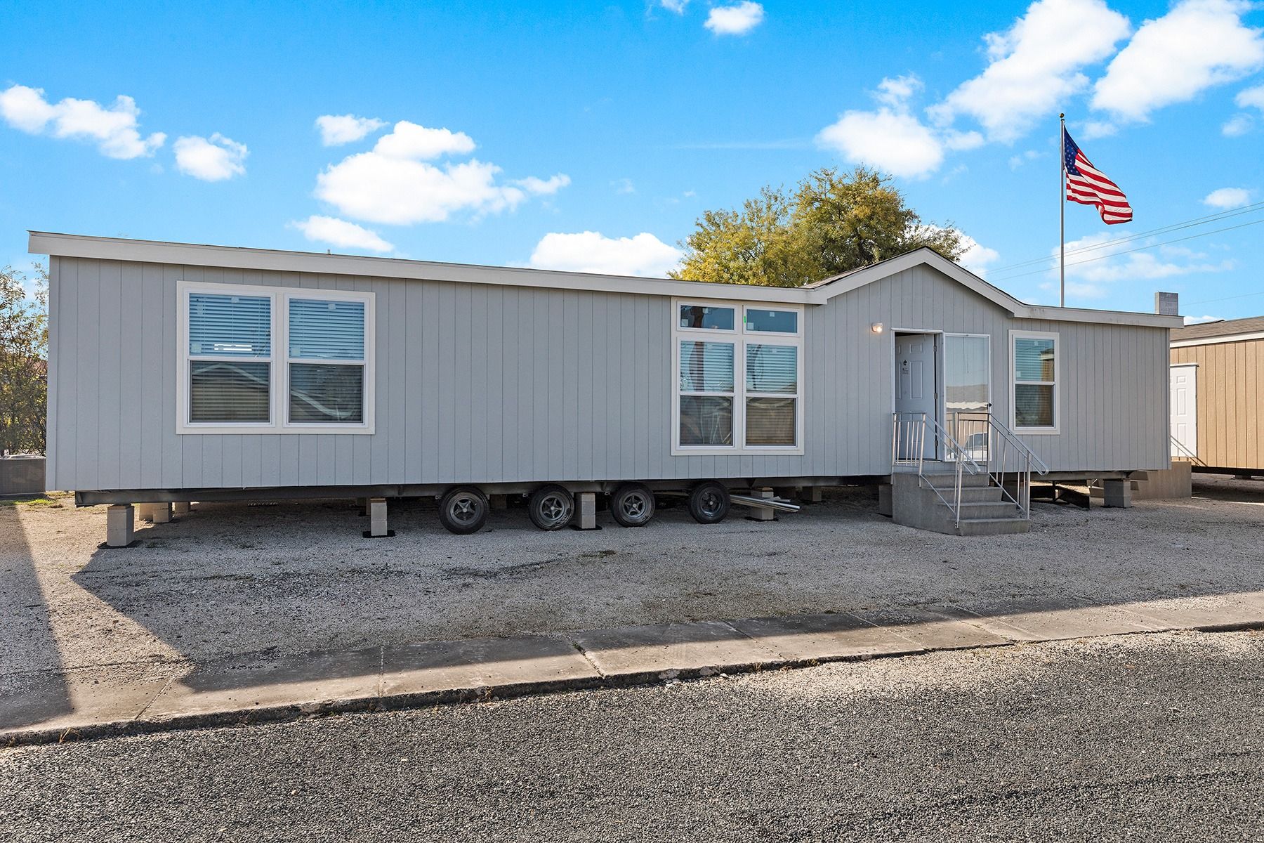 Gray mobile home with white trim on gravel lot under a blue sky, with an American flag nearby